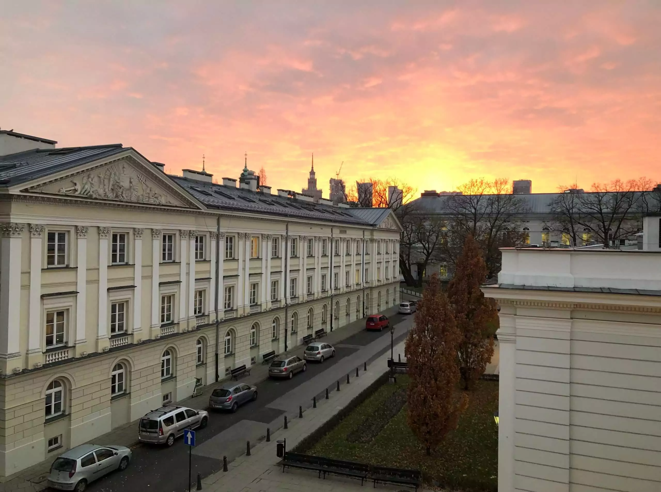 Widok na budynek Wydziału Prawa i Administracji, wewnętrzną uliczkę, na której stoi kilka samochodów, kilka drzew oraz w oddali widoczny jest budynek Wydziału Nauk Politycznych i Studiów Międzynarodowych. Na tle zachodzącego słońca widać budynki w centrum Warszawy